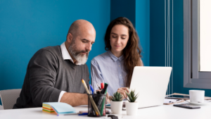 Two coworkers collaborate at a white desk, looking at an open laptop with pens, sticky notes, and small plants nearby in a blue office.