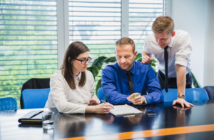 Three colleagues collaborate at a conference table, reviewing documents as one man points to notes on a pad while another watches; a window and plants are in the background.