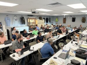 Group of adults in a classroom-style training session, dozens of laptops on tables, a ceiling projector, and gear displayed on the back wall.