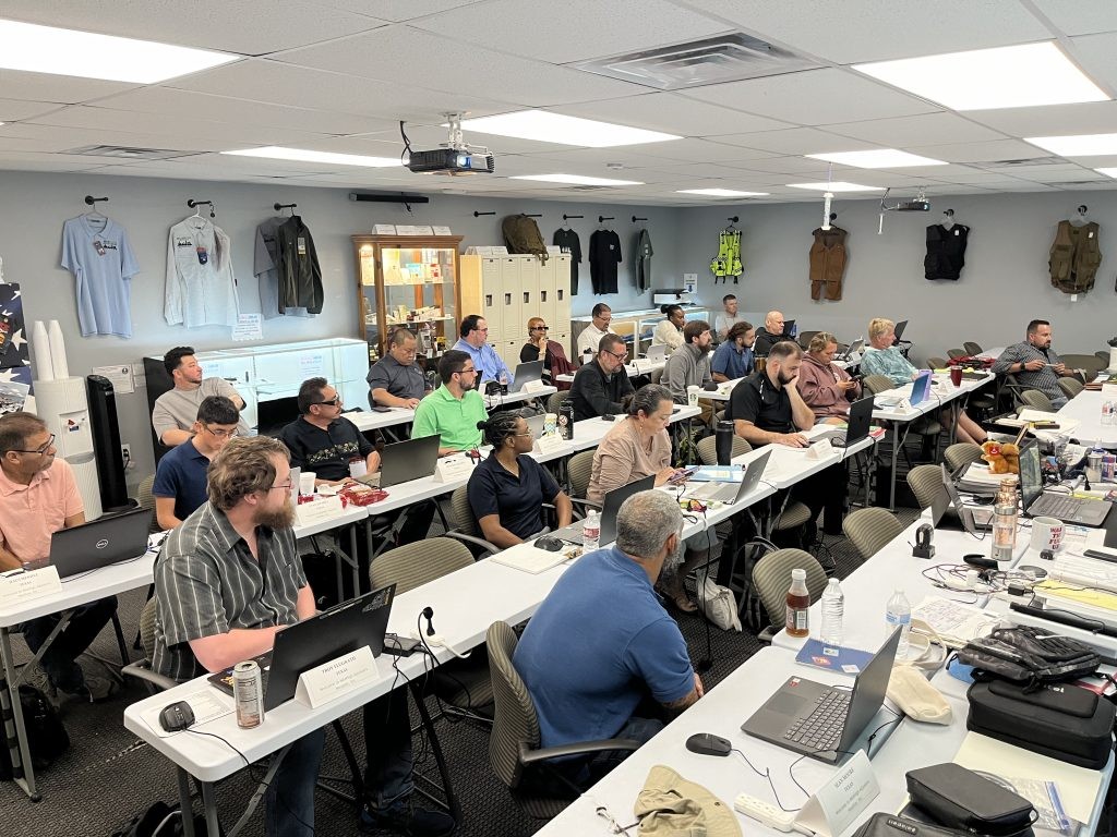 Group of adults in a classroom-style training session, dozens of laptops on tables, a ceiling projector, and gear displayed on the back wall.
